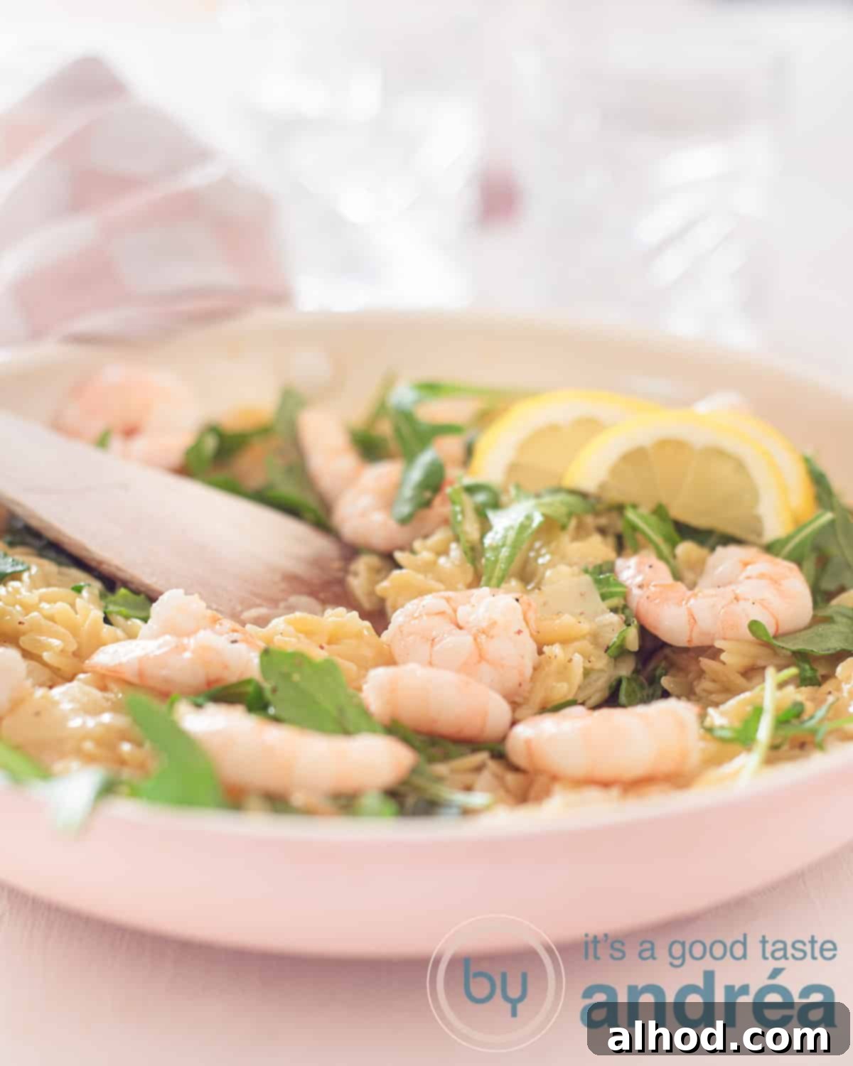 Zesty Lemon and Parmesan Arugula Risotto 5 An overhead shot of a pink frying pan filled with creamy risotto, shrimp, arugula, and lemon slices. A wooden spoon is resting in the pan.
