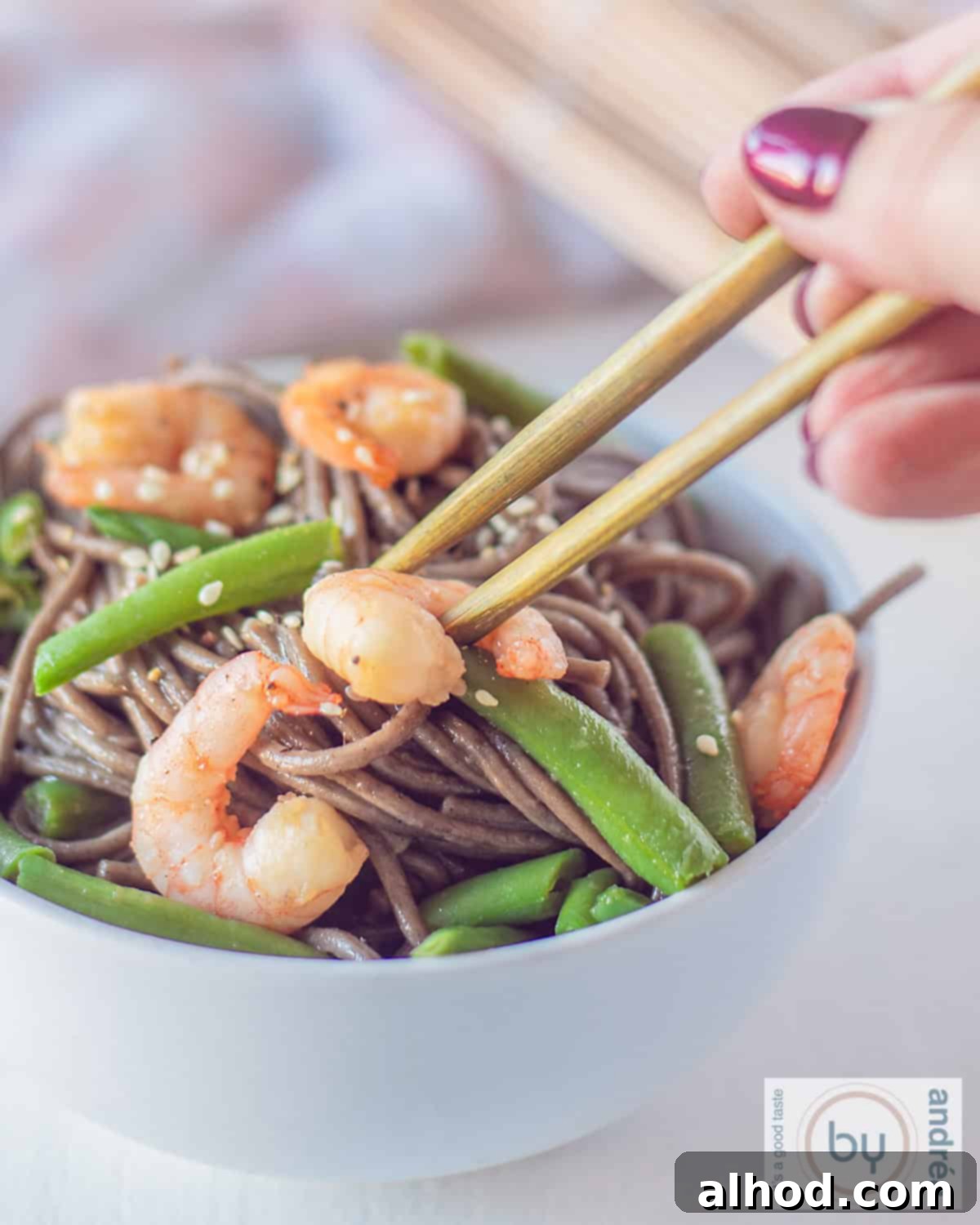 Shrimp and Green Bean Soba Ready in 20 Minutes 2 A pair of chopsticks picking up a juicy shrimp from a white bowl filled with green tea-infused soba noodles, bright green beans, and toasted sesame seeds, ready to be enjoyed.