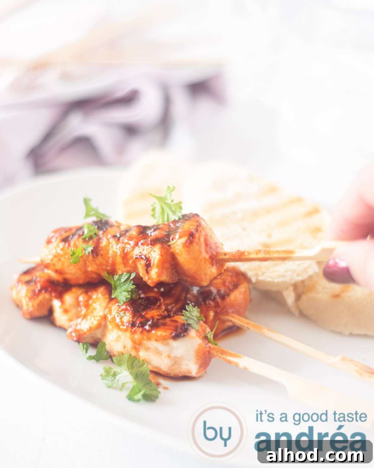 Bourbon Glazed Chicken and Bacon Skewers 2 A skewer with chicken with a bacon paste marinade on a white plate. A hand takes a kebab.