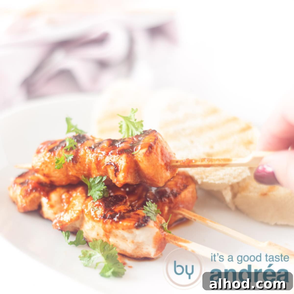 Bourbon Glazed Chicken and Bacon Skewers 4 A square photo with a white plate with three chicken skewers, a hand grabbing one and three slices of toast
