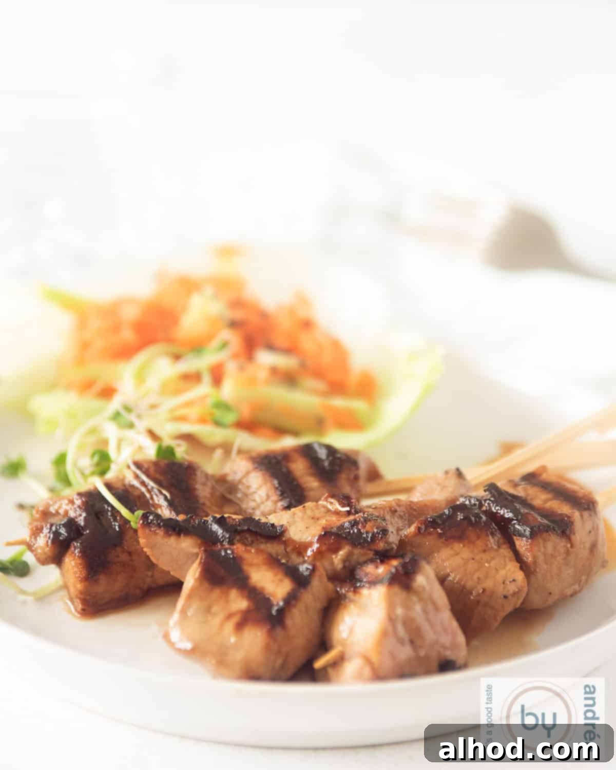 Sweet & Savory Asian Skewers with a Cooling Veggie Salad 2 An overhead shot of a white plate with three grilled pork tenderloin skewers and a lettuce leaf with cucumber carrot salad in the background.