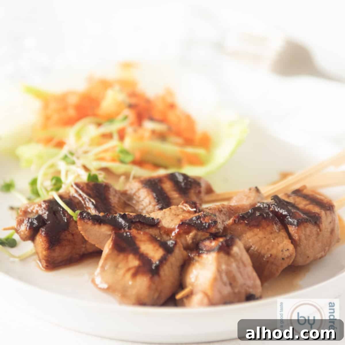 Sweet & Savory Asian Skewers with a Cooling Veggie Salad 4 A square photo of a white plate with three skewers of pork tenderloin with ketjap and a salad with carrot and cucumber in the background
