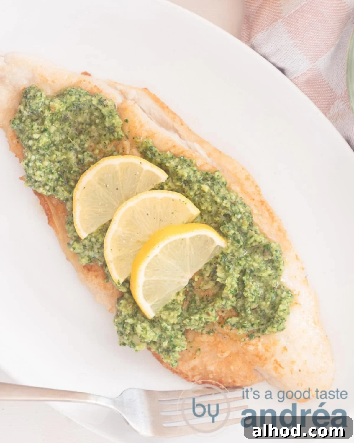 A photo from above of a pangasius fillet with pesto and lemon, garnished and ready to serve. At the top right a pink and white checkered tea towel adds a touch of charm.