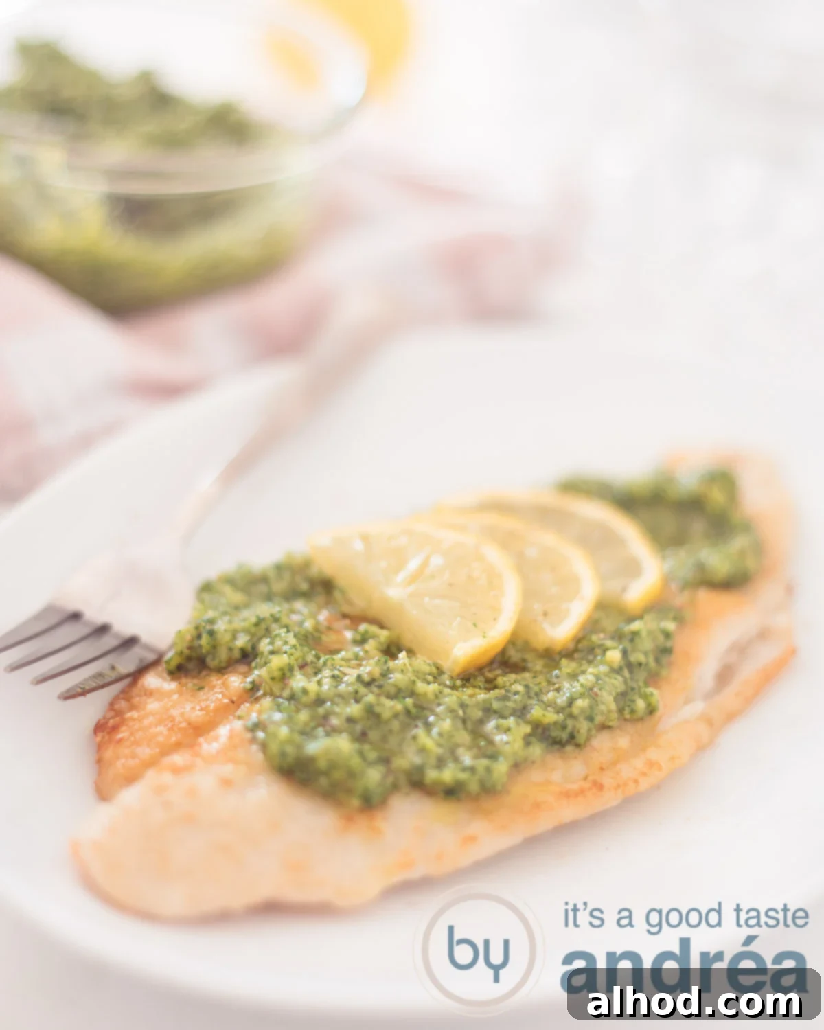 Bright Lemon Pesto Pangasius 5 A white plate with a pangasius fillet topped with arugula pesto and a slice of lemon in the foreground. In the background, a pink and white cloth and a glass bowl filled with arugula pesto.