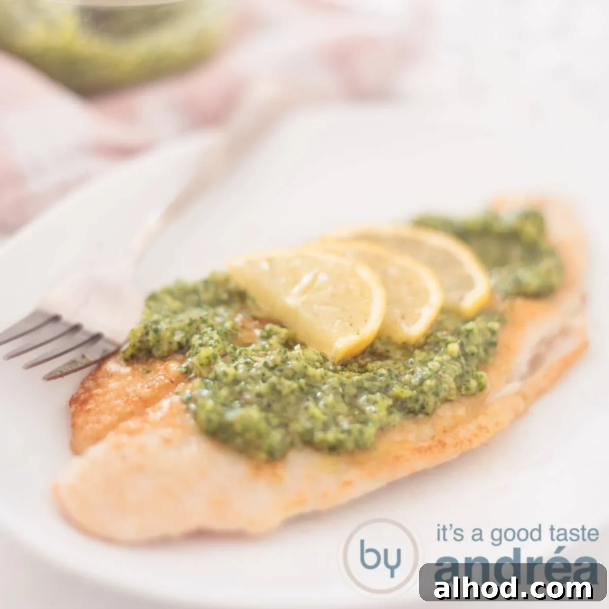 A square photograph showing part of a white plate with arugula pesto topped pangasius fillet with lemon slices, a vibrant and healthy meal.