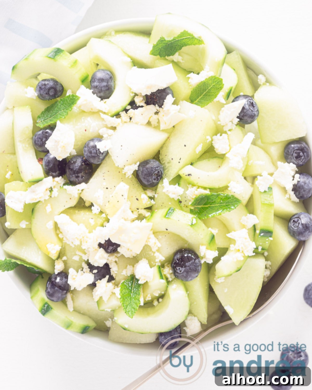 Melon Berry Bliss 2 A photo from above of a white bowl filled with diced melon, cucumber moons, blueberries, crumbled feta and a golden spoon. A white background