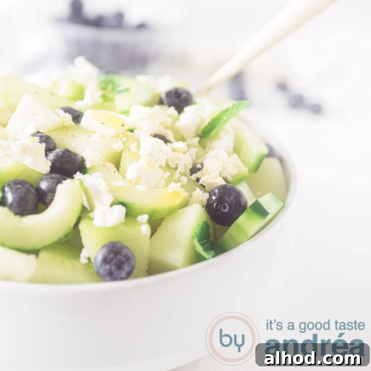 Melon Berry Bliss 6 A square photo showing part of a white bowl filled with blueberry melon salad with feta and cucumber on a white background.
