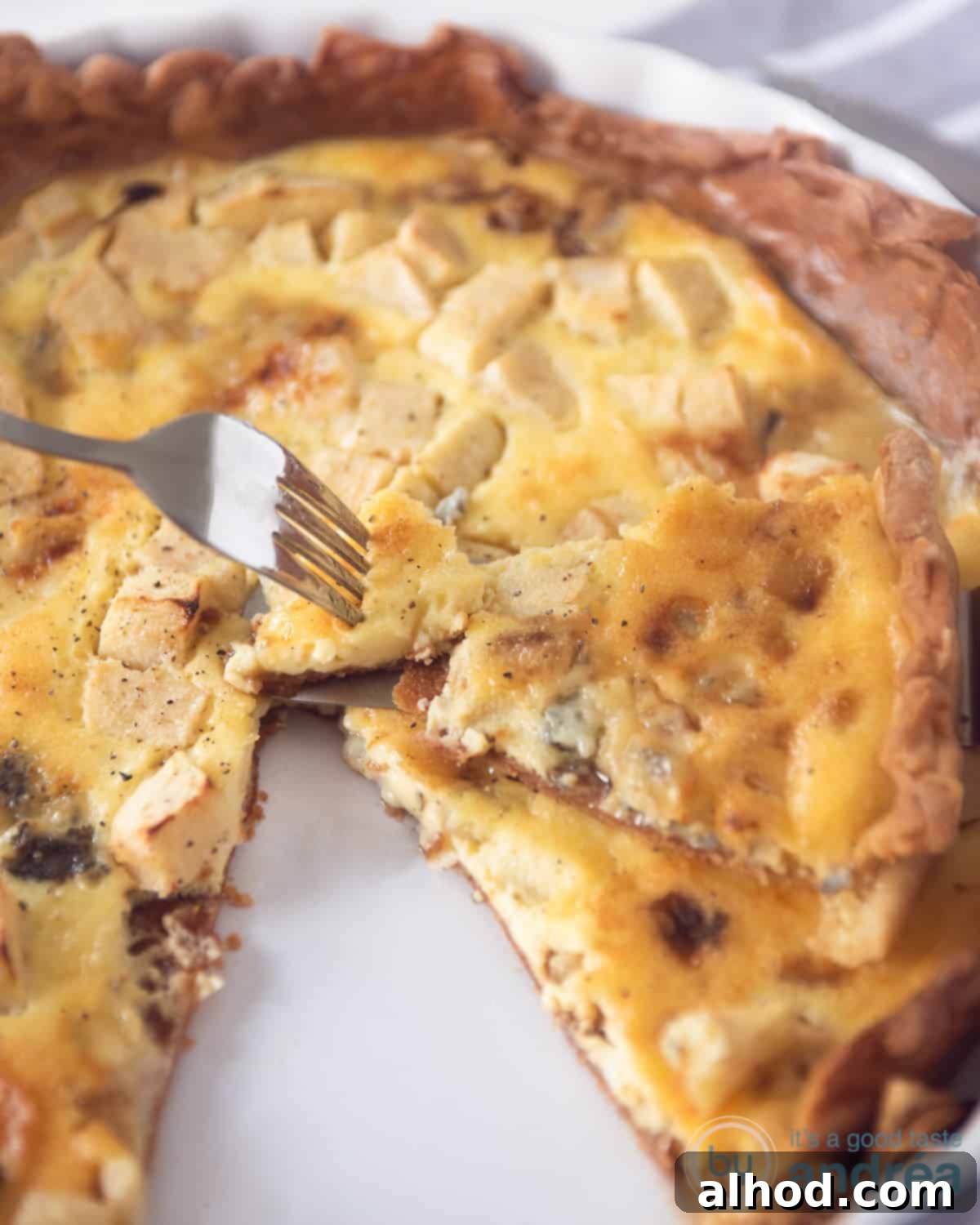 A high-angle shot of a Gorgonzola Apple Quiche with a slice removed, resting on top of the main pie. A fork is shown taking a bite from the quiche slice, highlighting its delicious texture.