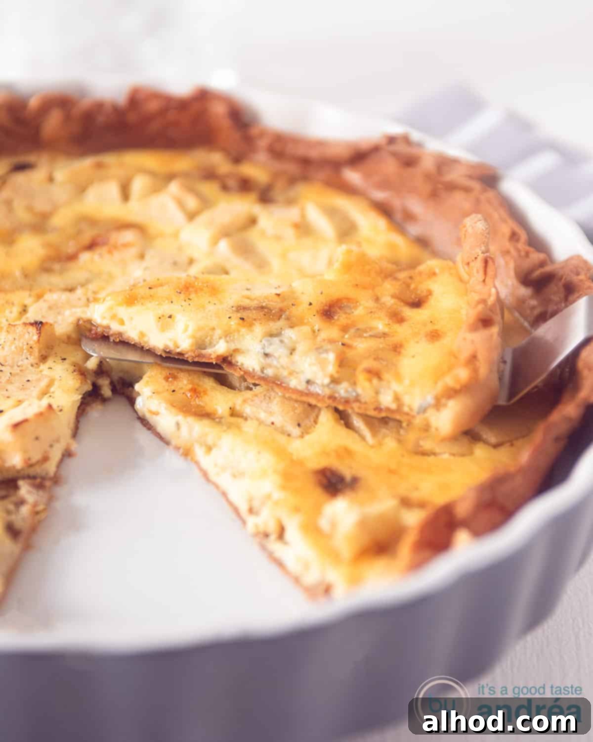 A grey ceramic quiche dish proudly holding a baked Gorgonzola and apple quiche. A perfect slice, removed from the main pie, rests enticingly on top of it, ready to be enjoyed.