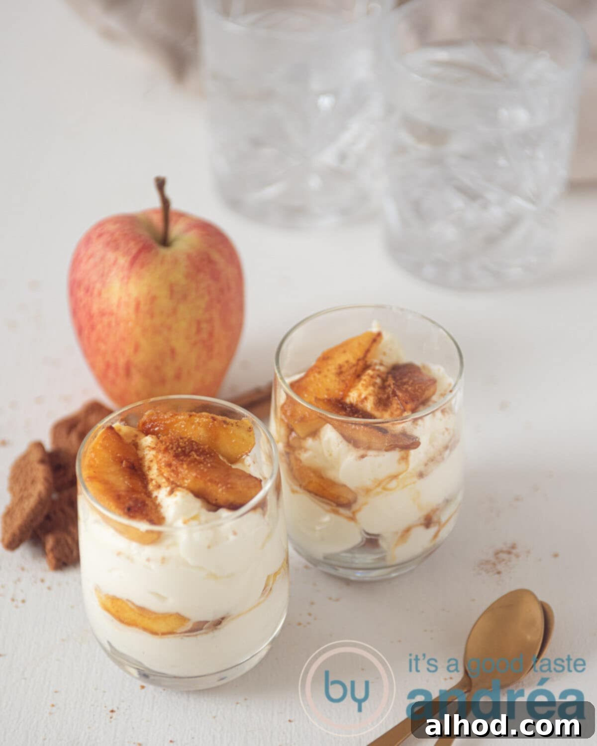 Two glasses from above, beautifully layered with whipped cream, caramelized apples, and a crushed cookie base. A fresh apple is in the background, and two spoons are in the foreground, ready to dig in.