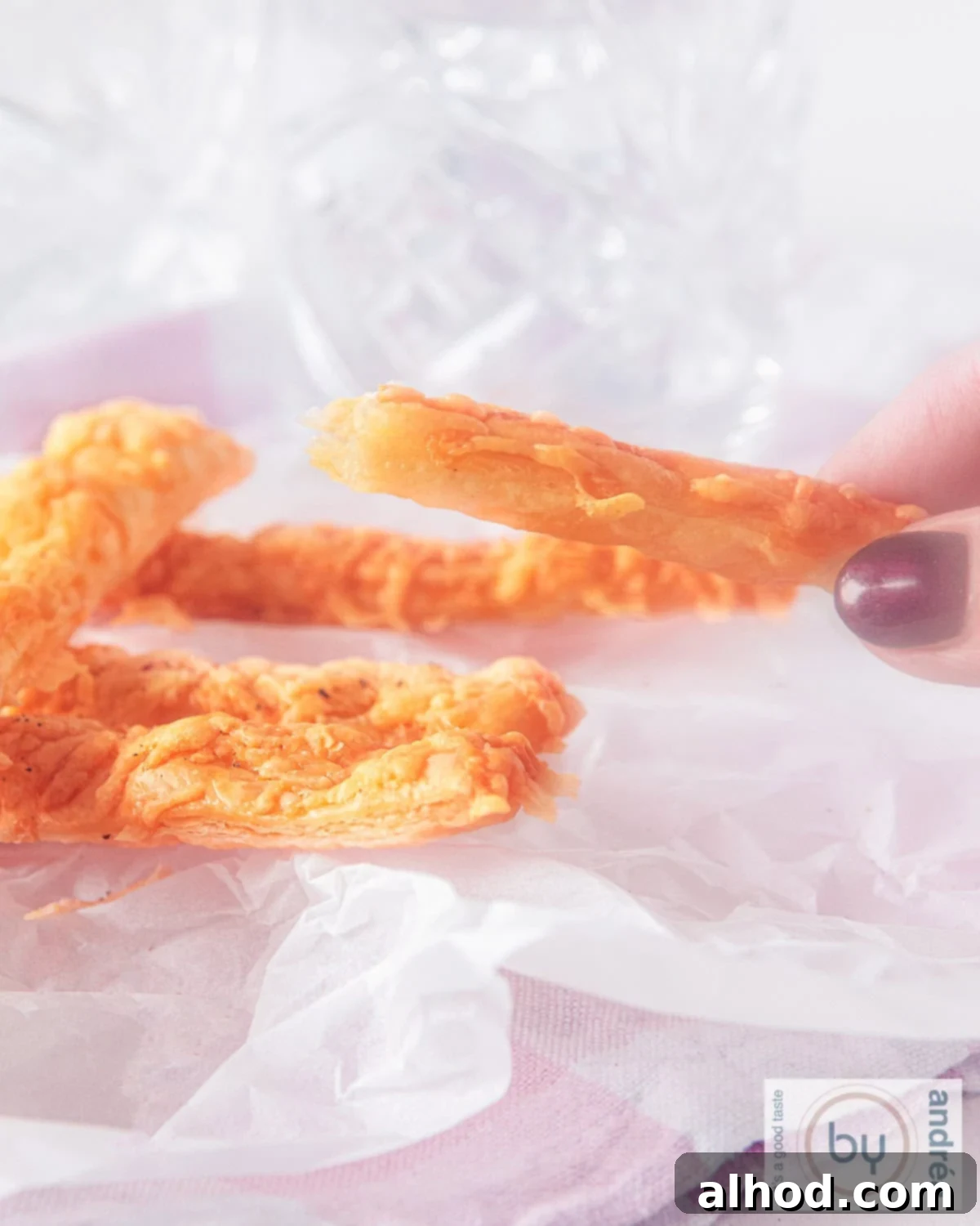 Easy 3 Ingredient Parmesan Puff Pastry Sticks 2 A height photo with a few cheese sticks on baking paper, one hand holding a stick.