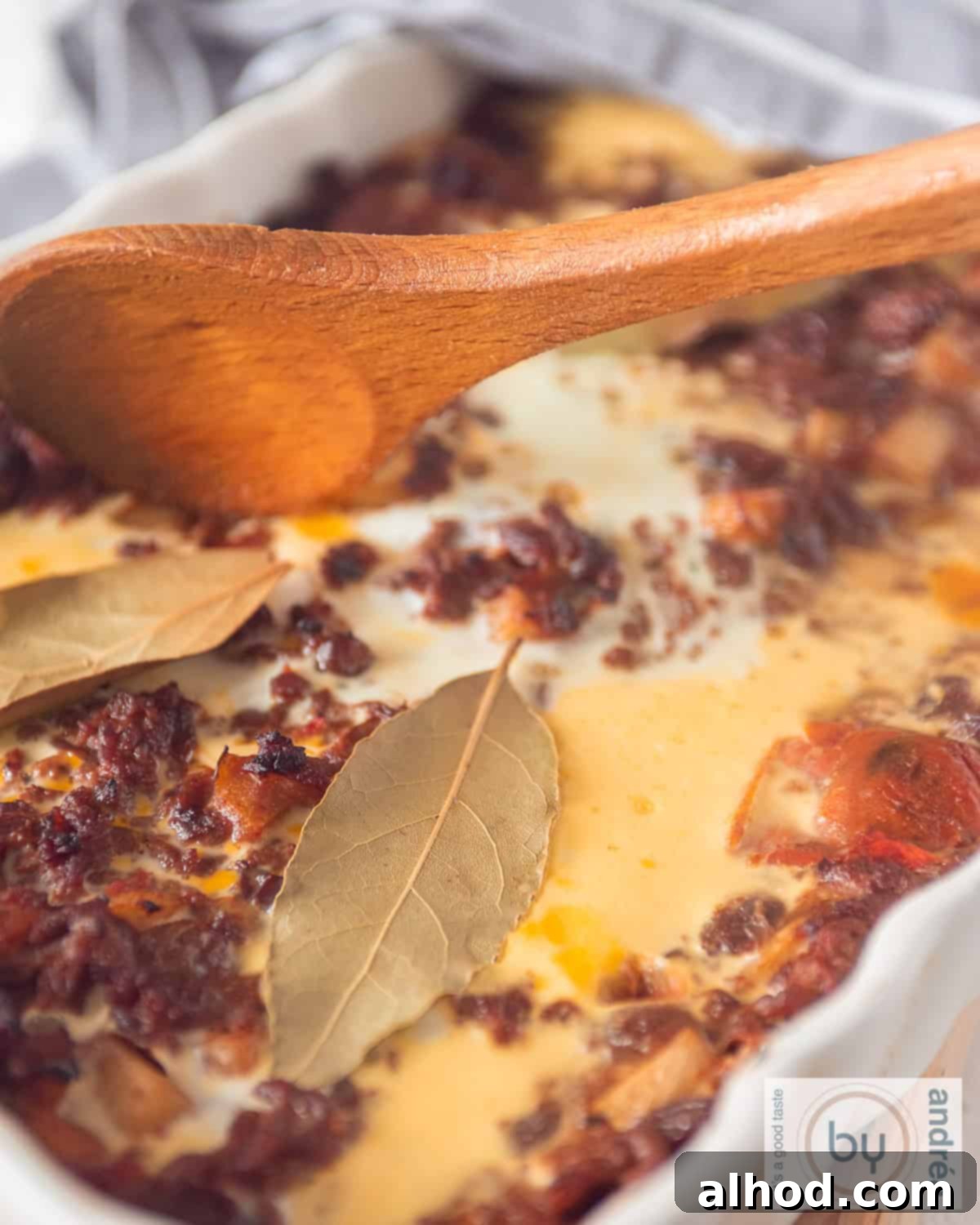 A portion of a white baking dish with an egg layer, minced meat, bay leaf, apple and raisins: African Bobotie