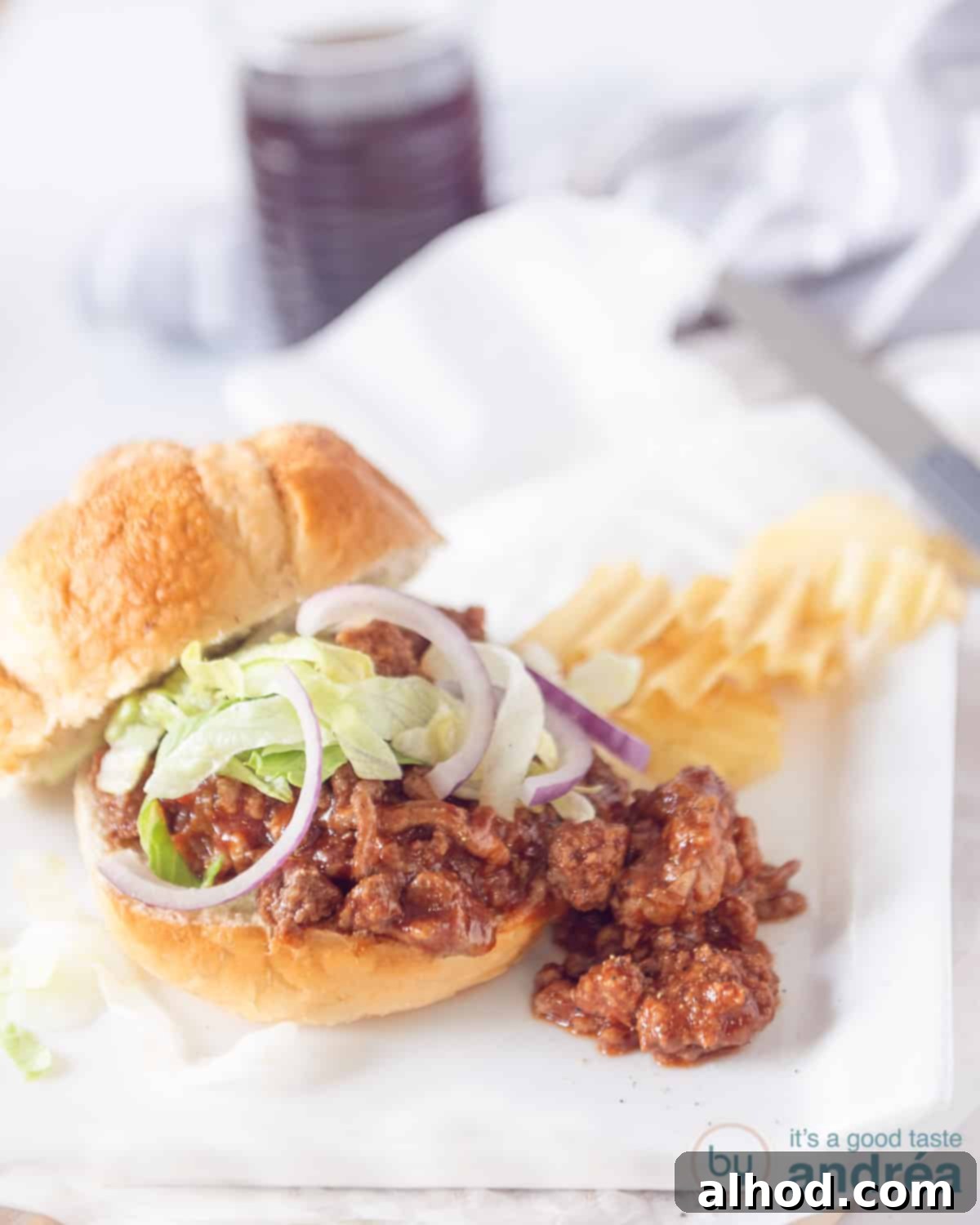 Effortless Slow Cooker Sloppy Joes 2 A perfectly assembled sloppy joe sandwich with a generous helping of minced meat, fresh lettuce, and crisp red onion. Ribbed potato chips and a glass of cola are blurred in the background, completing a classic American meal.