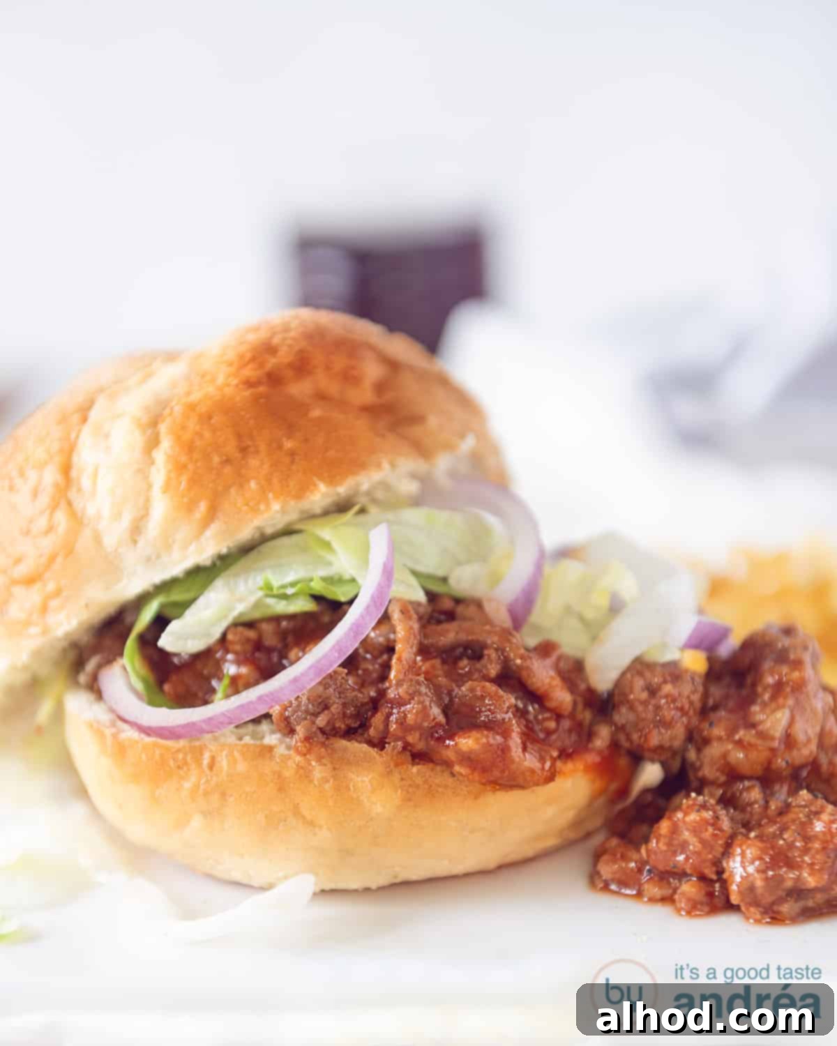Effortless Slow Cooker Sloppy Joes 3 A close-up of a delectable sloppy joe bun, generously filled with the savory meat mixture, topped with fresh red onion slices and vibrant green lettuce, all presented on a clean white background.