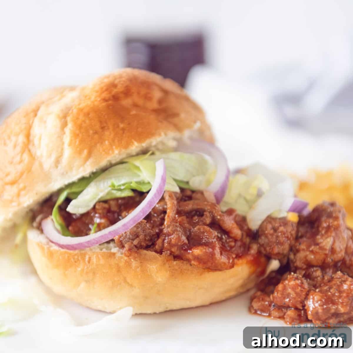 Effortless Slow Cooker Sloppy Joes 4 A square photo of a hearty sloppy joe sandwich, generously topped with savory minced meat in a rich sauce, garnished with crisp lettuce and vibrant red onion, ready to be enjoyed.