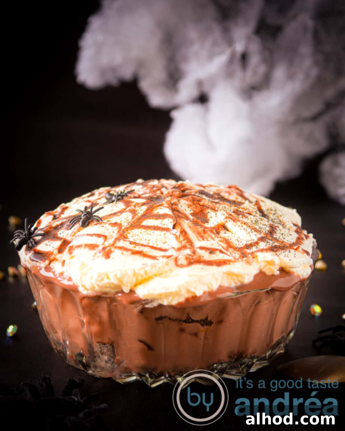 Oreo Brownie Graveyard Trifle 2 A glass bowl showcasing a delicious layered chocolate Oreo trifle. The top layer features whipped cream with a caramel spider web design, perfect for Halloween. The background is black, adorned with decorative spiders.