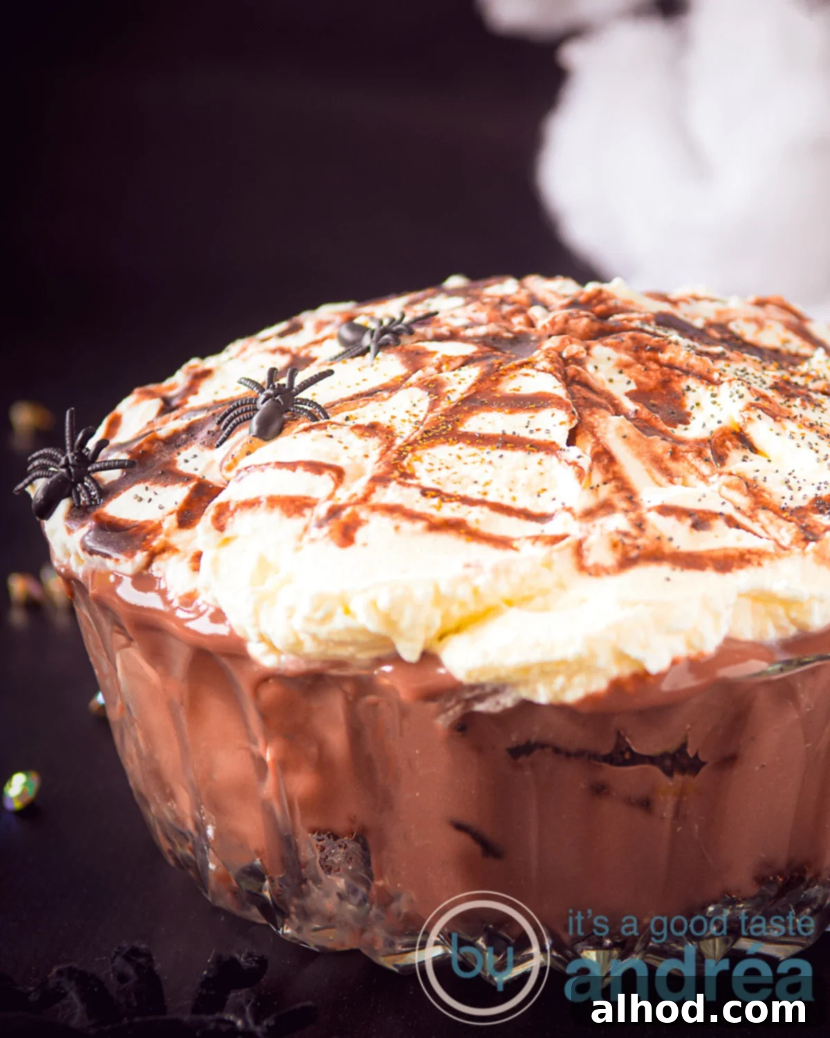Oreo Brownie Graveyard Trifle 4 A stunning close-up of a glass bowl filled with rich chocolate mousse, chunks of brownie, and crushed Oreo cookies, topped with a decorative caramel spiderweb. The dessert is ready to be served for Halloween.