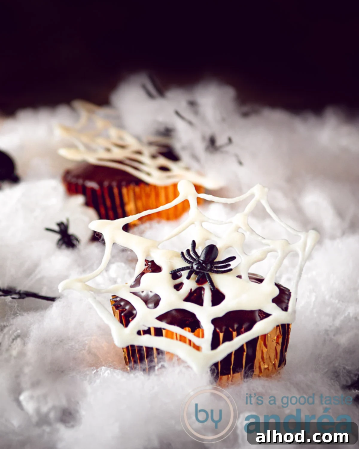 Spooky Nutella Chocolate Spiderweb Cupcakes 2 Three chocolate cupcakes with a white chocolate cobweb on top, set on a cloudy white surface with a dark background. Pin description: Delicious home-baked chocolate cupcakes, filled with Nutella and dipped in chocolate glaze. A white chocolate spider web cob completes this Halloween cupcake.