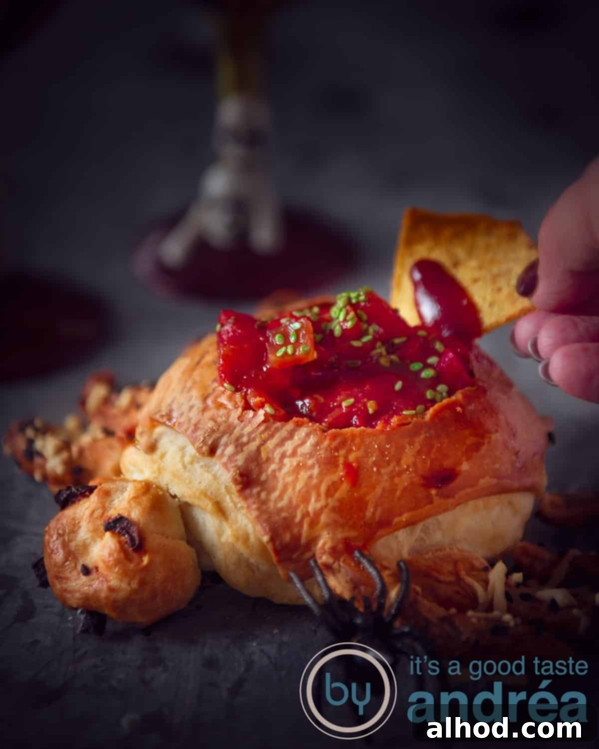 Frightfully Simple Halloween Nacho Dip 3 A hand holding a nacho dipping in the taco sauce contained in a bread spider shaped. A dark background