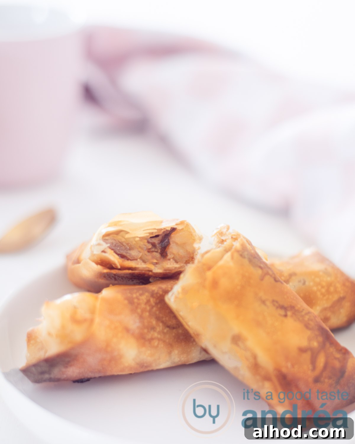 Apple turnovers with filodough. One whole on a white plate and two halves on top. You can see the filling. A pink cloth in the back.