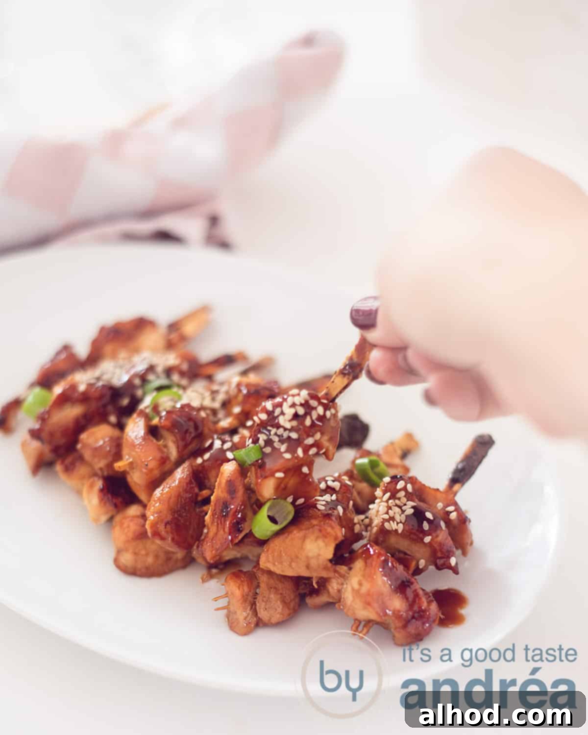 Authentic Japanese Chicken Yakitori 5 A white plate with about 20 Japanese chicken skewers with yakitori sauce, sesame seeds or spring onion. A hand taking a skewer. A white background