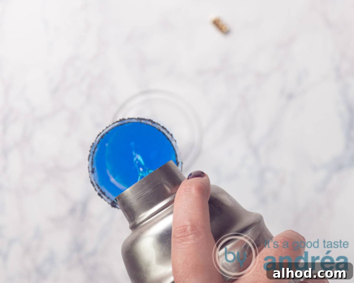 Pouring a vibrant blue Halloween cocktail through a sieve into a glass with a black sugar rim, ensuring a smooth, ice-free drink.