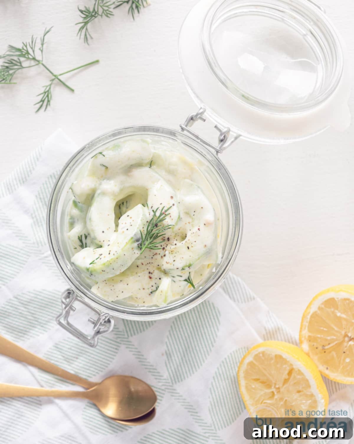 Refreshing Cucumber Yogurt Dip 2 A photo of a glass jar from above filled with Greek cucumber salad. Two lemon halves at the front right. Left for two golden spoons, inviting you to taste this fresh tzatziki.