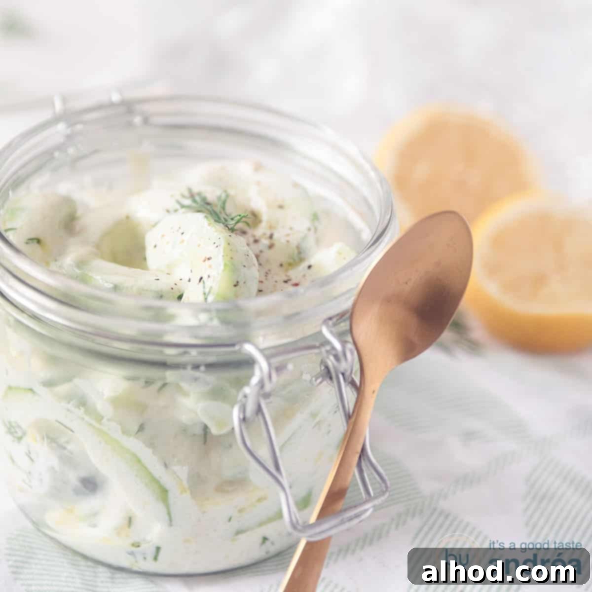 Refreshing Cucumber Yogurt Dip 5 A square photo with a glass jar containing a cucumber salad with Greek yogurt. There's a spoon in there for it. Two lemons in the top right.