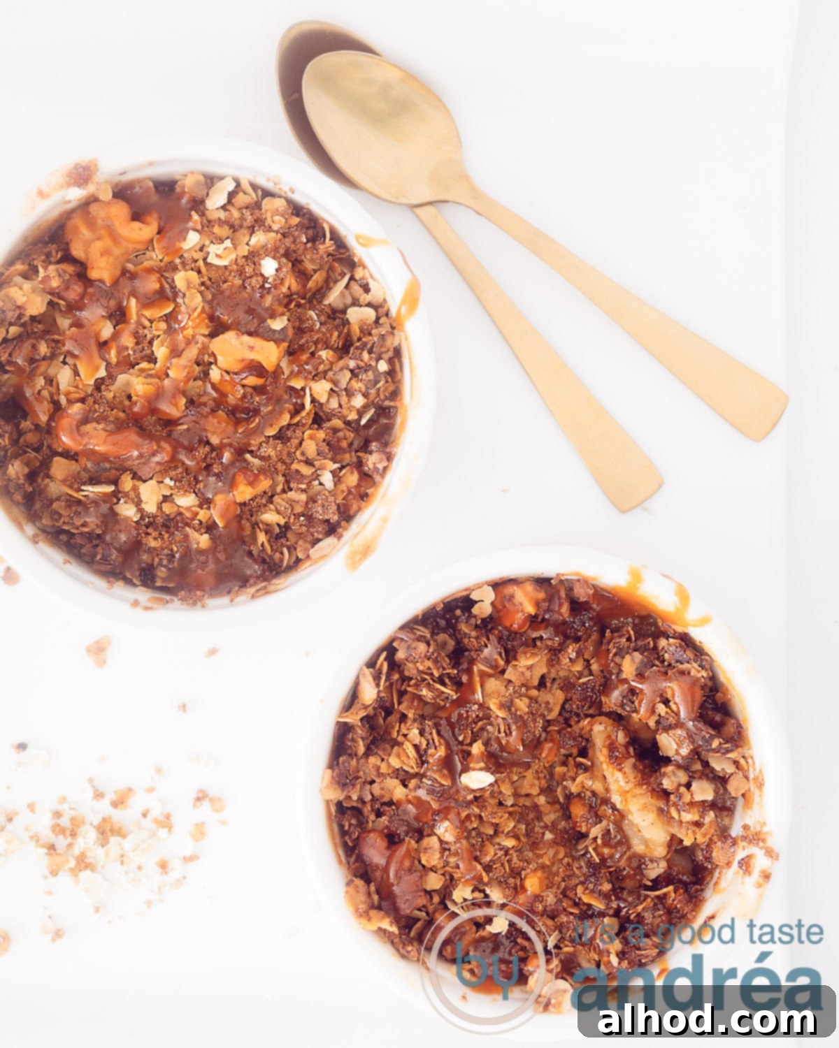 A photo from above of two white bowls with stewed apple and baked granola. On a white background, at the top right two golden spoons