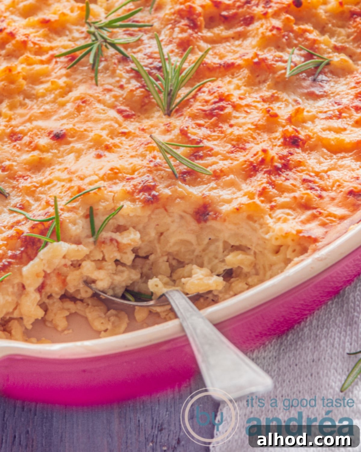 Homestyle Baked Mac and Cheese 2 A small portion of a pink baking dish filled with mac N Cheese. A spoon has taken a bite out of it. Rosemary lies on top of the garnished cheese.