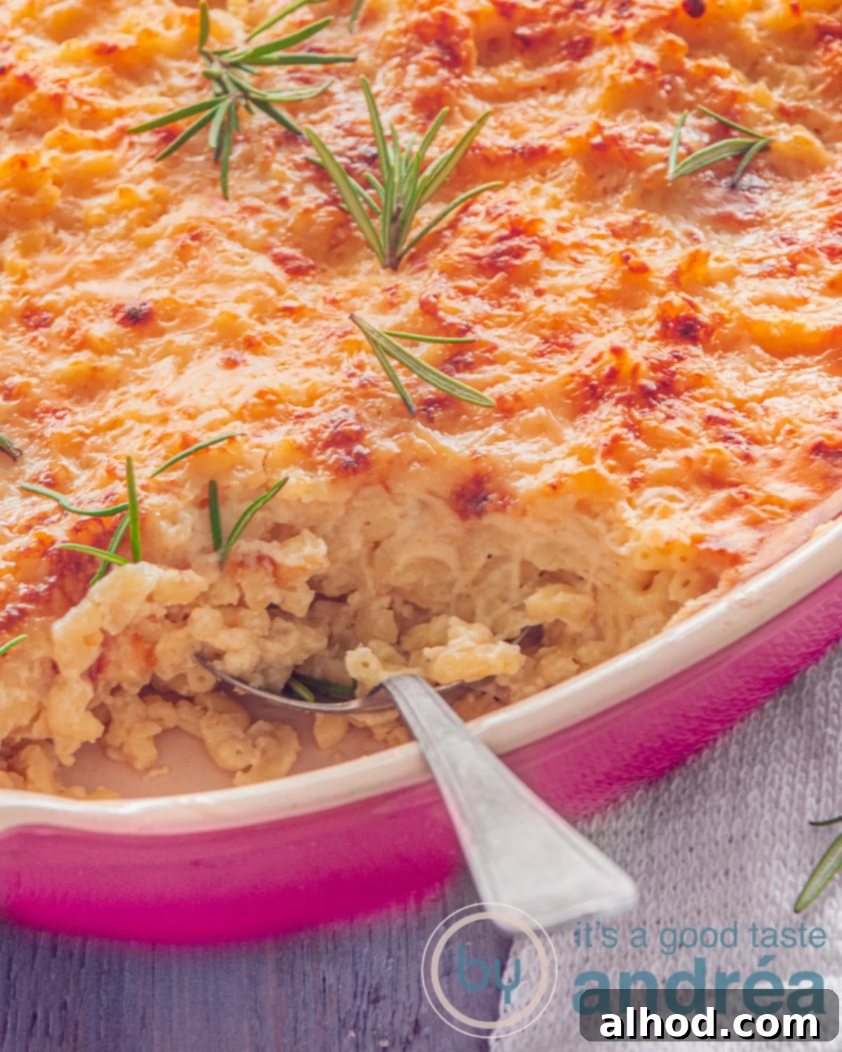 Golden Baked Mac and Cheese 2 A small portion of a pink baking dish filled with mac N Cheese. A spoon has taken a bite out of it. Rosemary lies on top of the garnished cheese, highlighting the rich, golden crust and creamy interior.