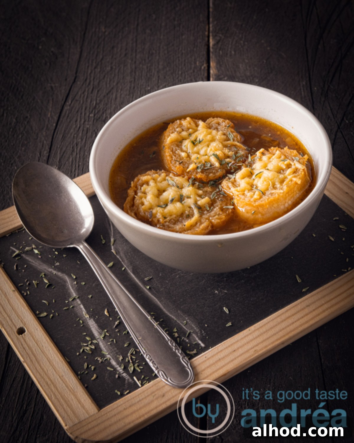 Savory Vegetarian French Onion Soup 5 A black background with a plate on it. On top a bowl with onion soup with baguette gratin and a spoon on the left.