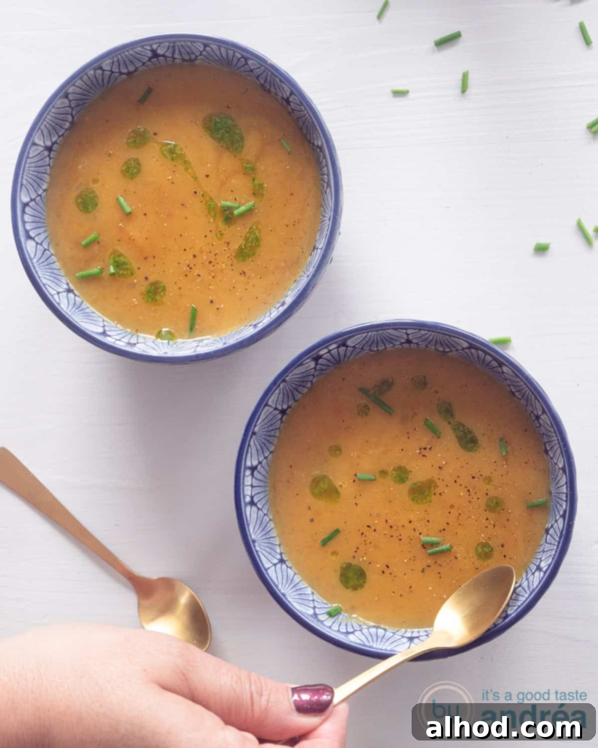 A photo from above of two blue soup bowls filled with roasted pumpkin soup, generously drizzled with vibrant green chive oil. A hand gently scoops a spoonful from the first bowl.