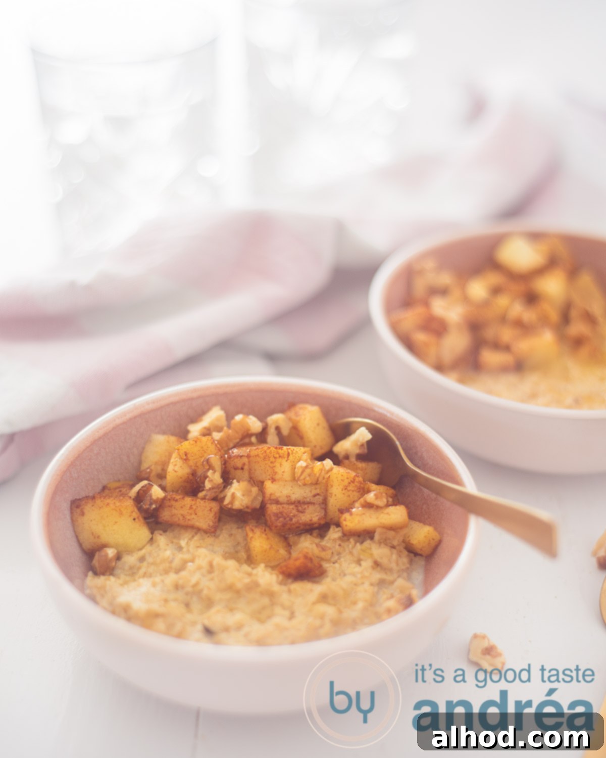 Warm Apple Cinnamon Oats 2 Two bowls with oatmeal, apple and cinnamon diagonally placed on a white background. A white pink cloth with two glasses in the back.
