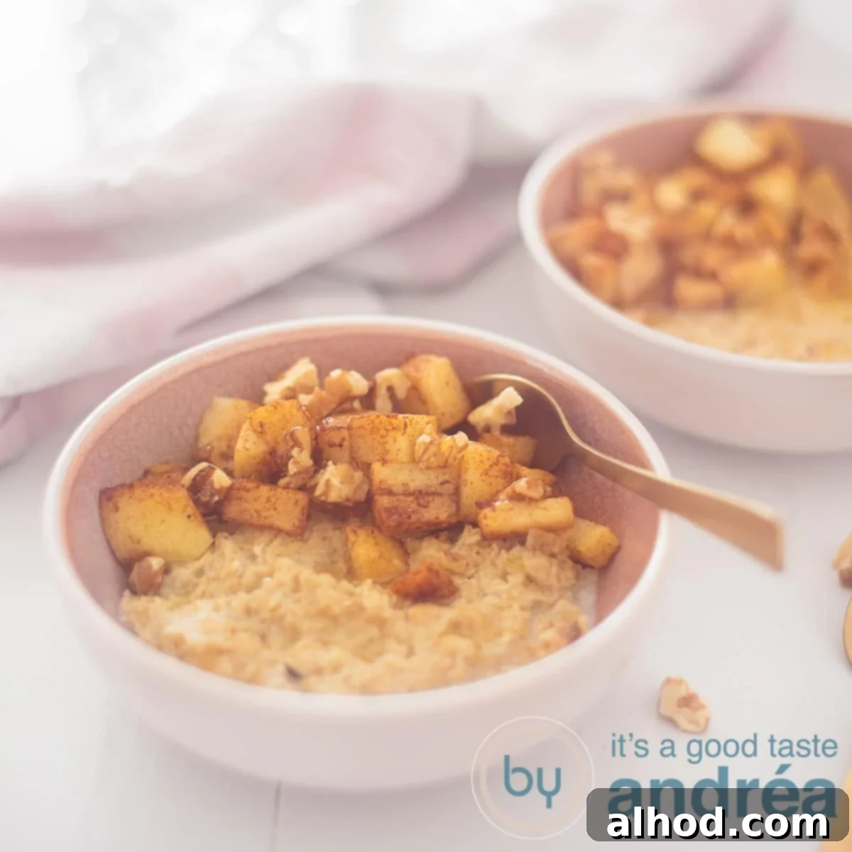 Warm Apple Cinnamon Oats 4 A square photo with a pink bowl in the foreground with oatmeal, apple, walnuts and honey. A golden spoon sticks into it. In the back right a second bowl. A white background with a pink and white tea towel at the top left.