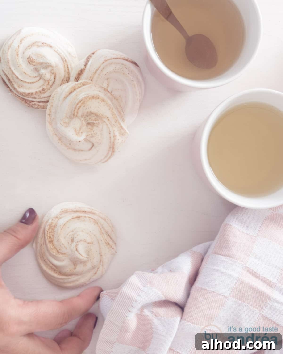 Snowy Kisses 2 A photo from above of four gingerbread meringues, one of which is being handled. Two mugs of tea on the right.