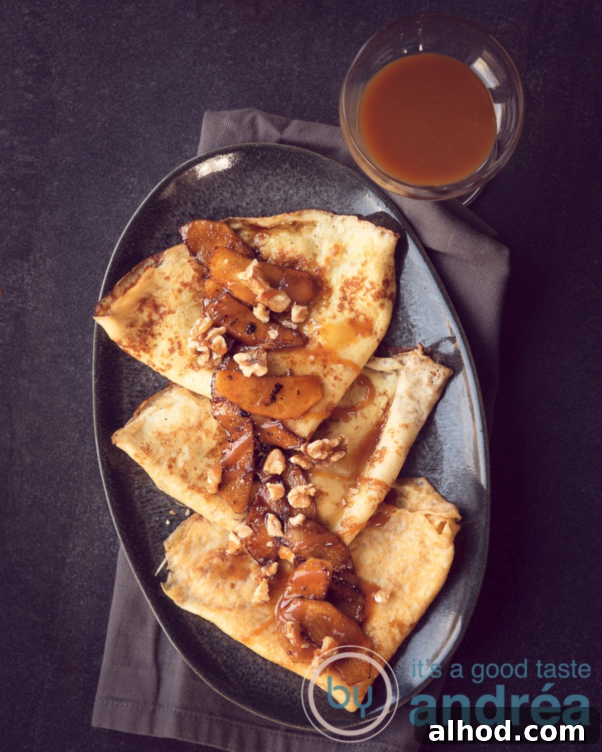 Raclette Sweet Treat: Caramelized Apple Crêpes 5 A grey platter holding three perfectly assembled crêpes, generously topped with caramelized apples, a rich cascade of caramel sauce, and a scattering of crunchy walnuts. A small bowl of extra caramel sauce is visible in the top right corner, all resting on a dark grey background.