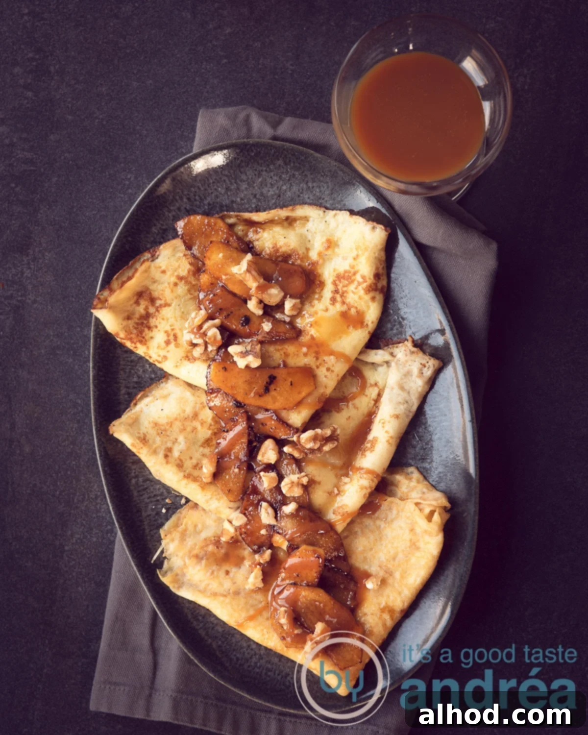 a grey platter with three crepes with apples, caramel sauce and walnuts. A bowl of caramel sauce on the right top. A dark grey background