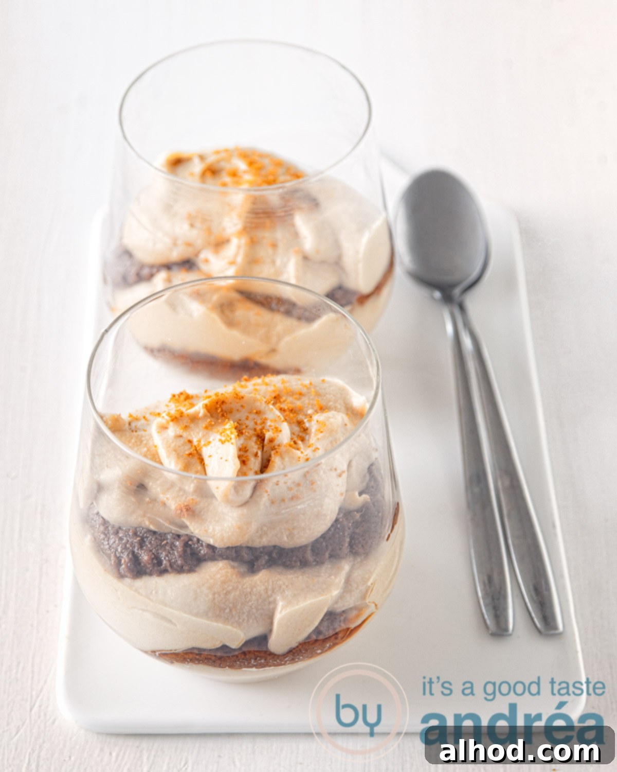 Creamy Biscoff Tiramisu 4 Two elegant glasses brimming with layered Biscoff Cookie butter cream and speculoos cookies, arranged on a pristine white plate, with two spoons invitingly placed beside them. This image highlights the delightful presentation of the dessert.