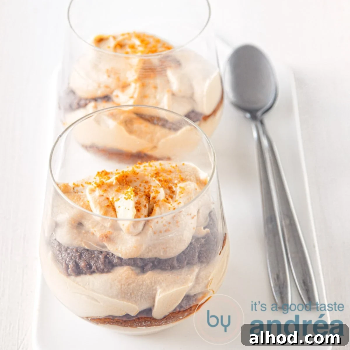 Creamy Biscoff Tiramisu 5 A square photo with two glasses of speculoos tiramisu behind each other on a white plate. Two spoons are next to it on the right.