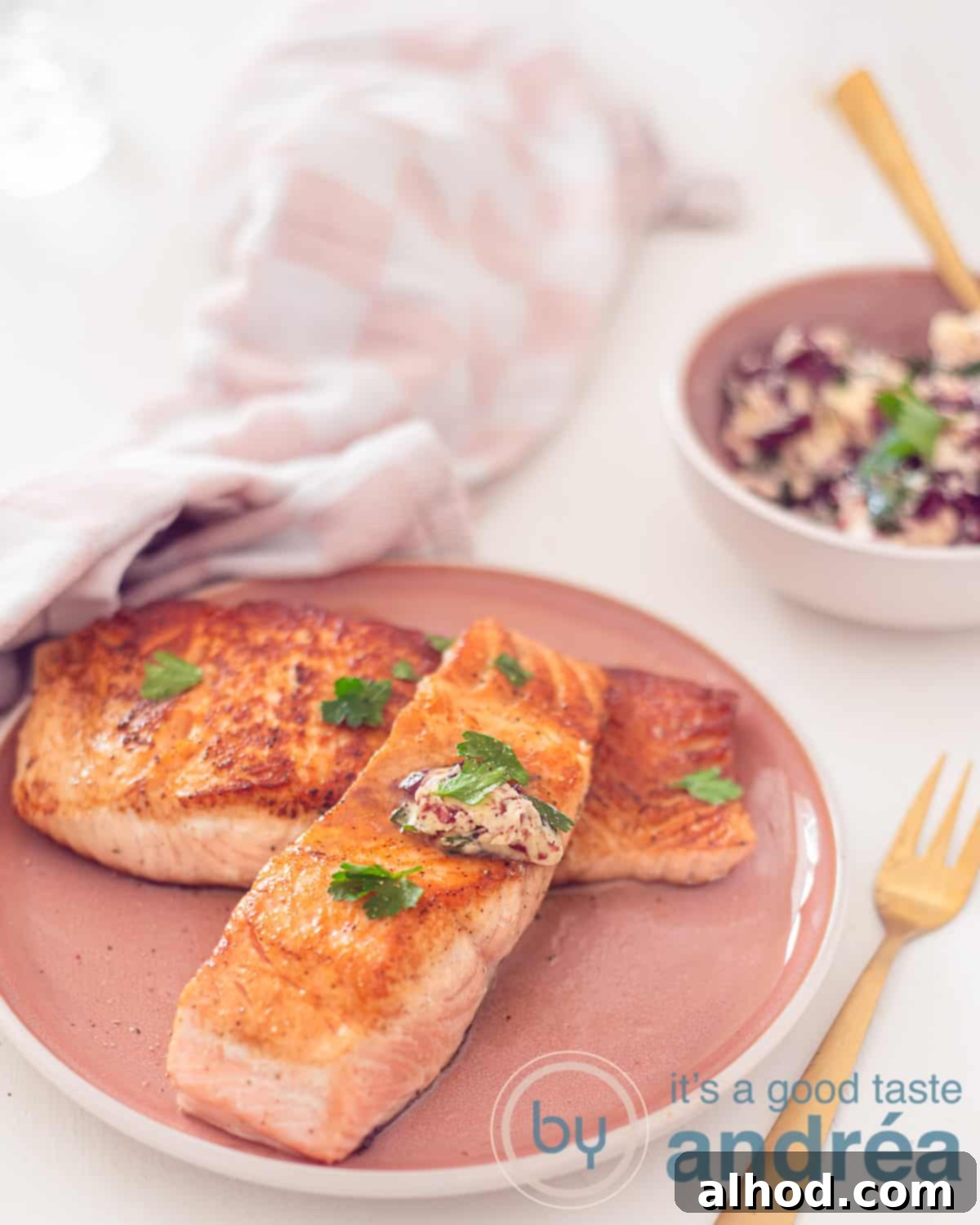 Two perfectly pan-fried salmon fillet steaks, generously topped with melted red wine herb butter, served on an elegant pink plate. A separate pink dish holds additional red wine herb butter, all set against a soft white background with a pink and white tea towel.