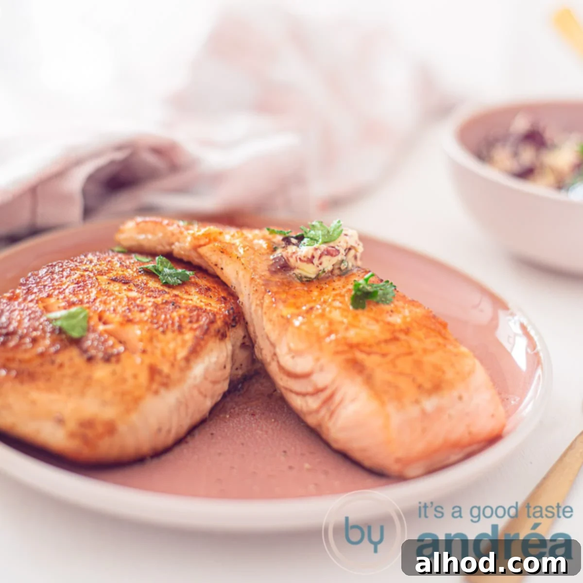 A square photo of a pink plate on a white background. Two baked salmon fillets with red wine herb butter on the plate. In the background a pink and white tea towel.