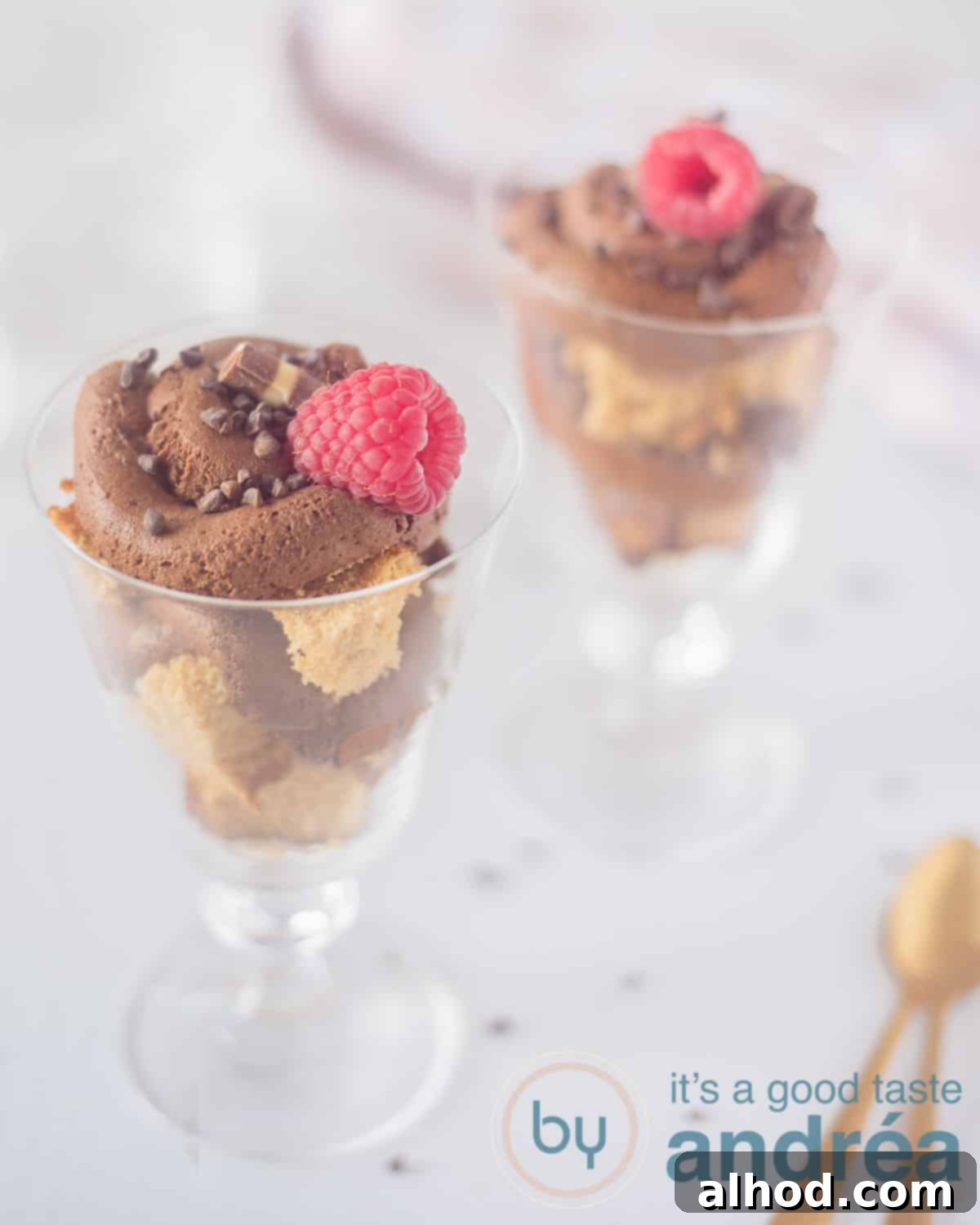 Decadent Chocolate Mousse Trifle 2 A height photo with two glass coupes filled with layers of cake alternated with chocolate mousse and top with a raspberry and chocolate. A white background. Bottom right two golden spoons.