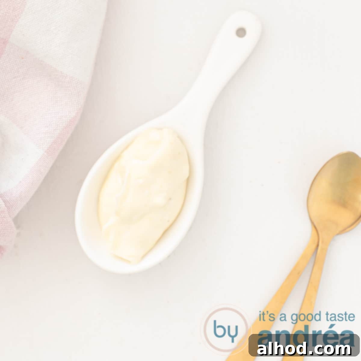 A square photo with a spoon of wasabi mayonnaise, two golden spoons on the side and a pink checkered tea towel in the top right, white background.