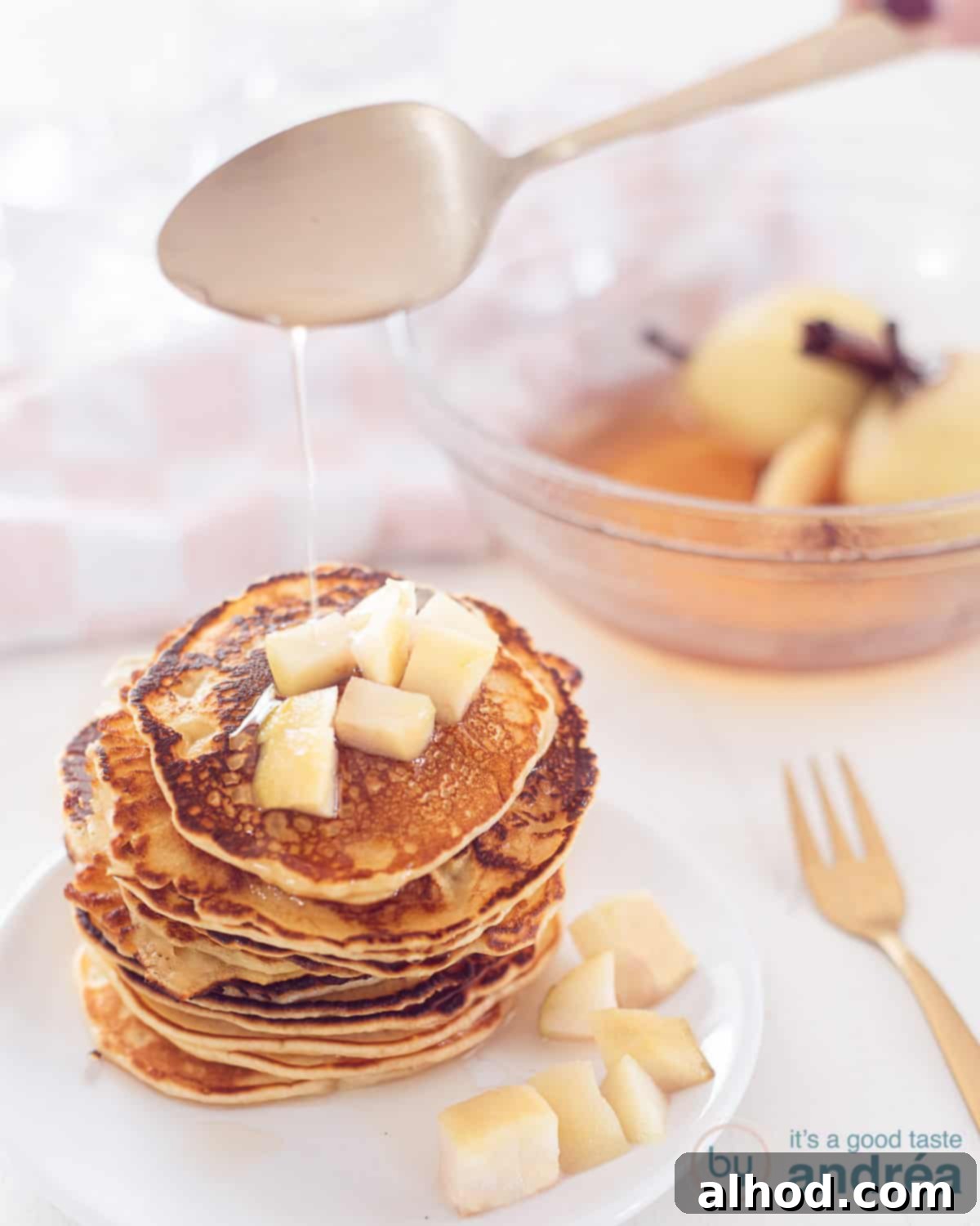 Poached Pear Pancakes 2 A white plate with a stack of pancakes with stewed pears. A golden spoon sprinkles some sauce on top. At the back right is a bowl with extra stewed pears and sauce.