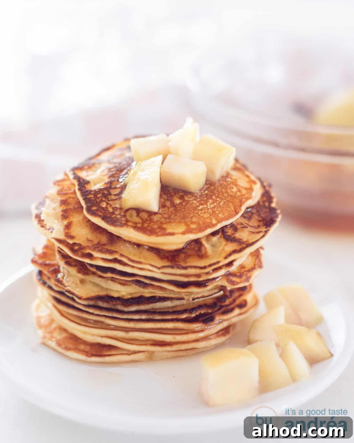 Poached Pear Pancakes 3 A stack of pancakes with poached pears, topped and around on a white plate. In the background a glass bowl with extra poached pears and syrup.