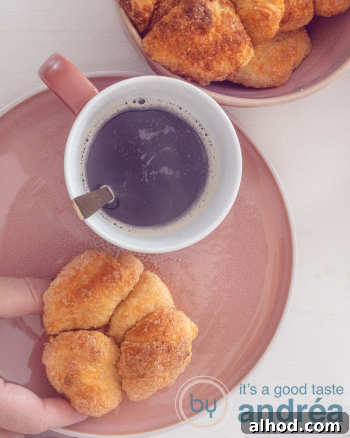 Pull-Apart Monkey Bread Muffins Recipe 2 A photo from above of a pink plate with a monkey bread muffin being taken off the plate. A cup of coffee next to it. Top right a bowl with extra muffins.