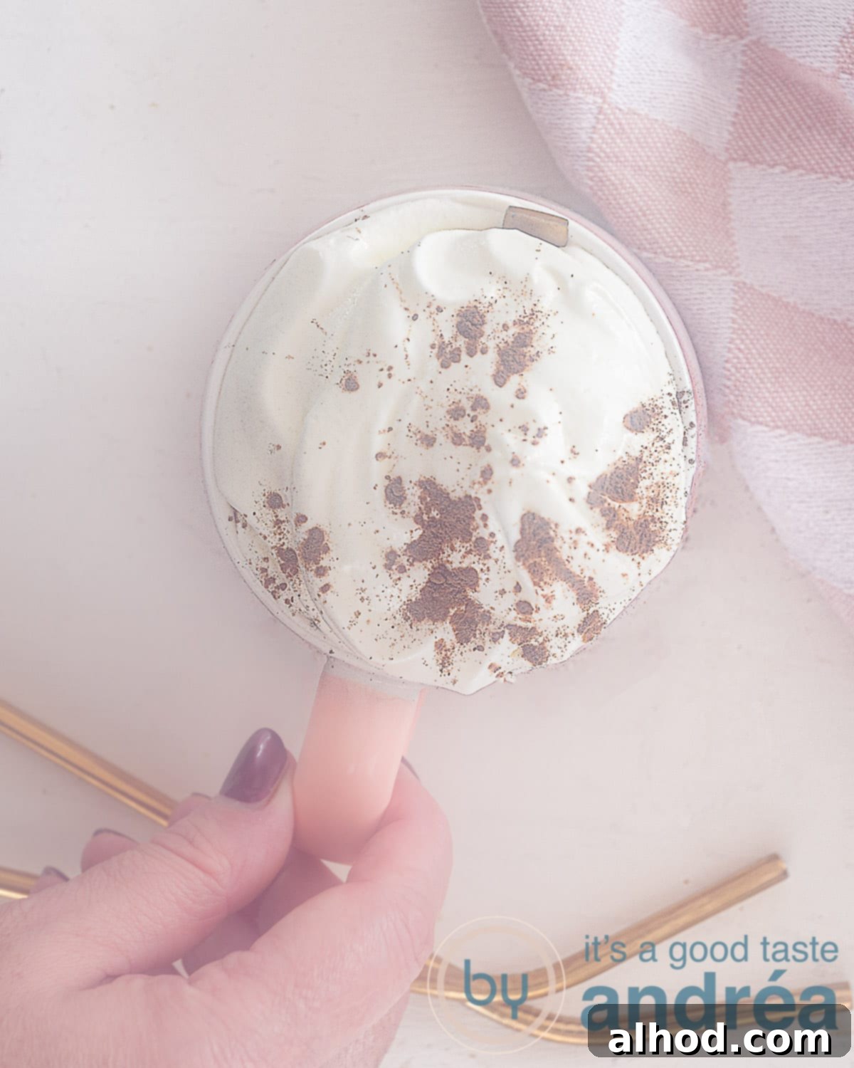 Decadent Baileys Hot Chocolate 2 A photo from above of a hand holding a mug of hot chocolate with Baileys. a white background. Top right a piece of a pink and white tea towel.