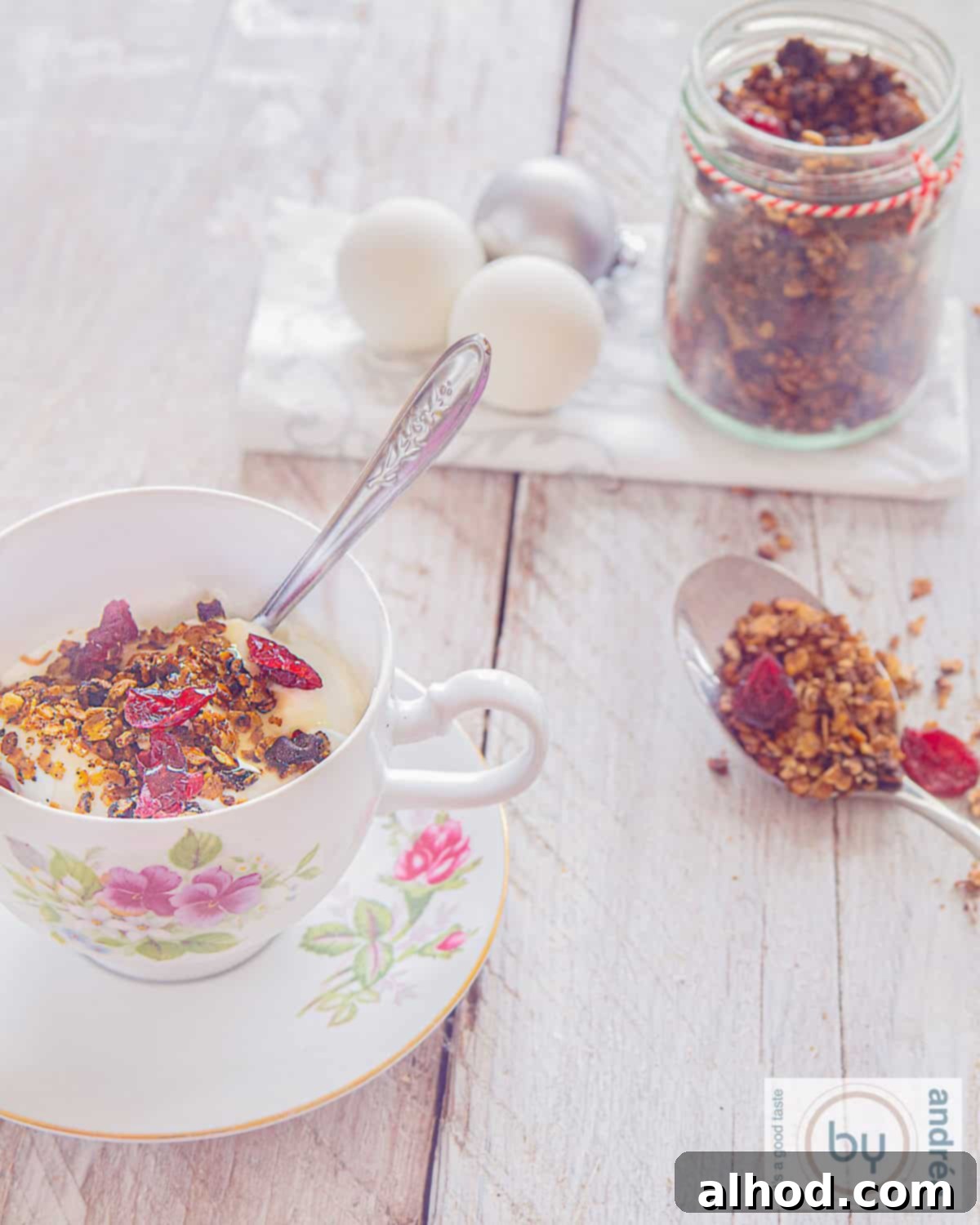 Yuletide Cranberry Pecan Crunch 3 A beautifully arranged breakfast setting featuring a cup of warm Christmas granola with yogurt and fresh cranberries. A decorative glass jar filled with extra granola and a spoon are visible in the background, highlighting its homemade appeal.