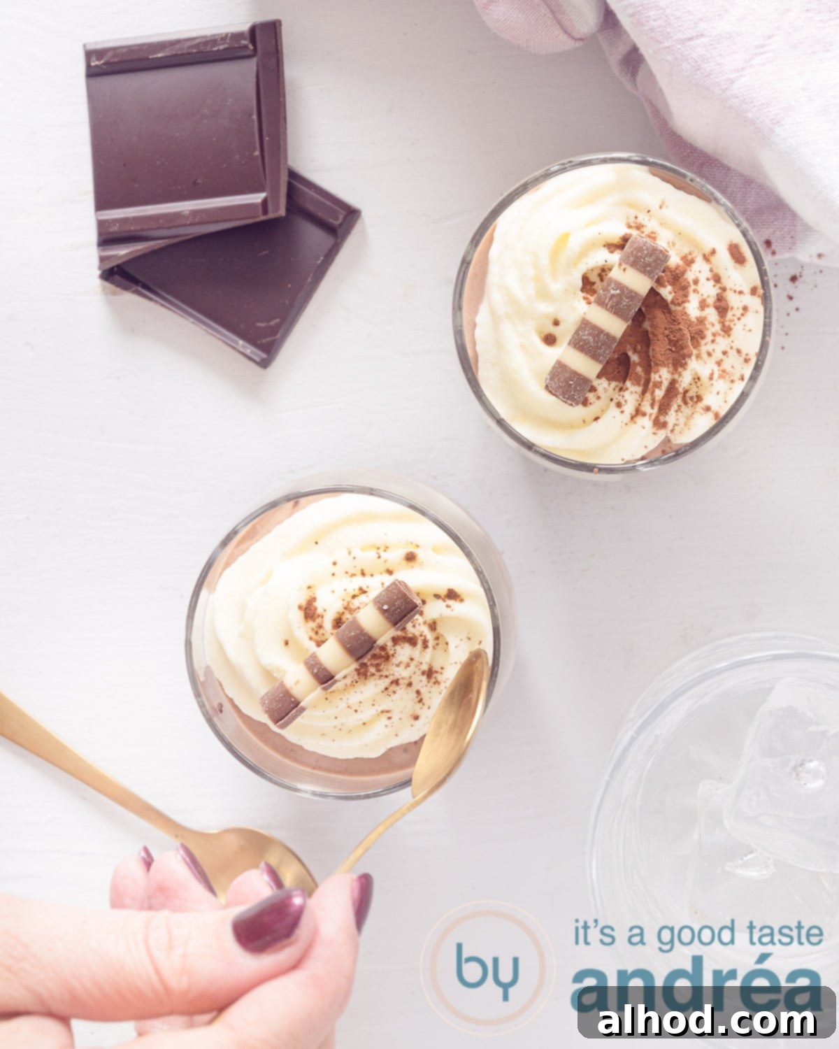 Decadent Triple Chocolate Mousse in a Glass 2 A photo from above of two glasses showing white chocolate mousse, two cubes of chocolate and a white background. A hand picks up a shovel.